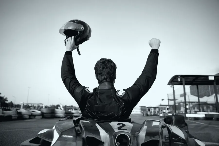 Piloto de costas, de pé no kart, erguendo o capacete com os dois braços.