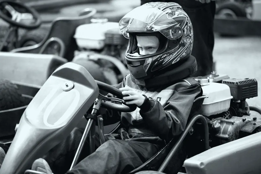 Criança de capacete sentada no kart, mãos no volante, em foto preto e branco.