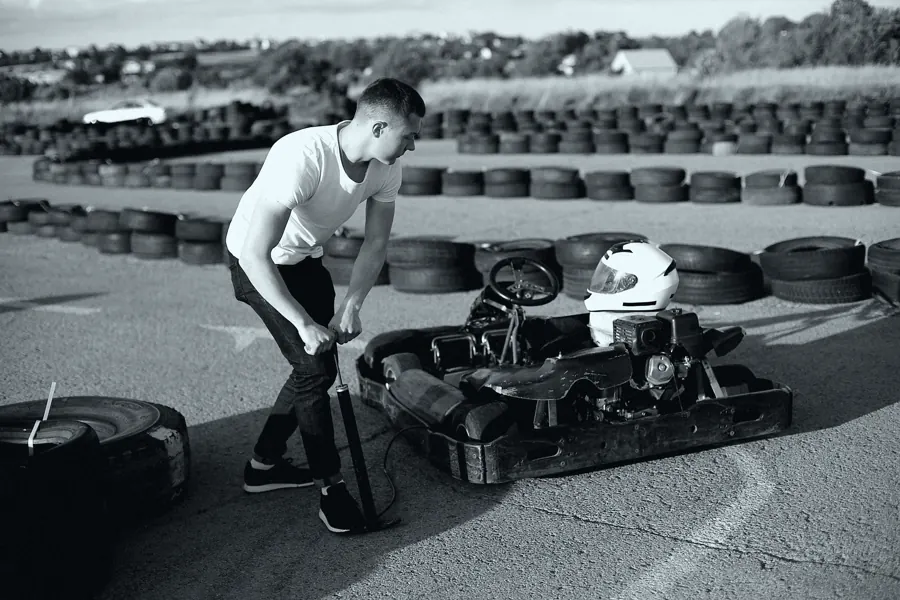 Mecânico de camiseta branca ajustando o kart ao lado da pista, com um capacete apoiado no banco.